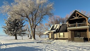 A snow-covered landscape with a wooden house featuring a balcony and a garage. - Newly renovated Ashton Farmhouse on Cave  Falls HWY. Near Yellowstone Nat. Park (Ashton)