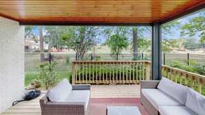 Un patio techado con dos sofás de mimbre y una pequeña mesa, con vista a un jardín cercado con árboles y una obra en construcción a lo lejos.