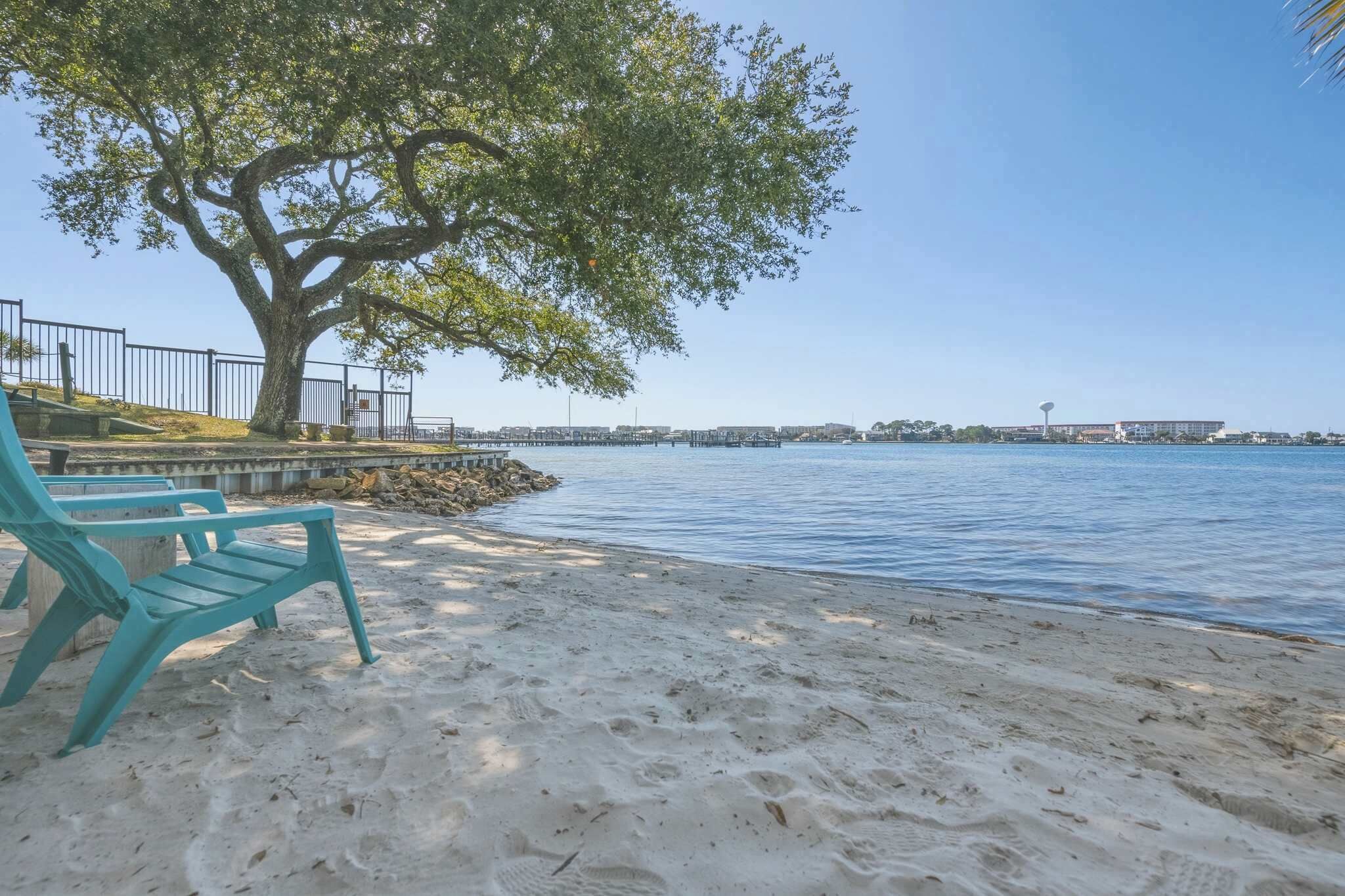 A sandy beach with a large tree and a row of turquoise chairs along the water's edge.