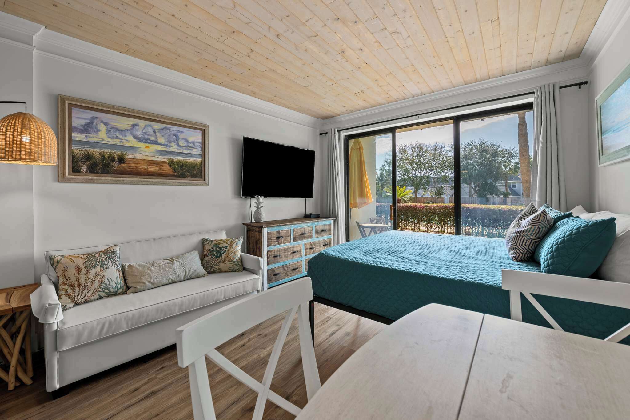 Een kamer met een houten plafond, een bank, een bed met blauwgroene beddengoed, een televisie, een raam met gordijnen en een schilderij aan de muur.