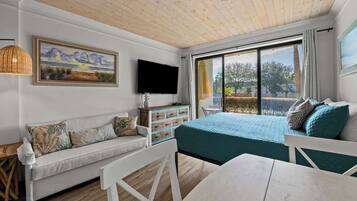 Een kamer met een houten plafond, een bank, een bed met blauwgroene beddengoed, een televisie, een raam met gordijnen en een schilderij aan de muur.