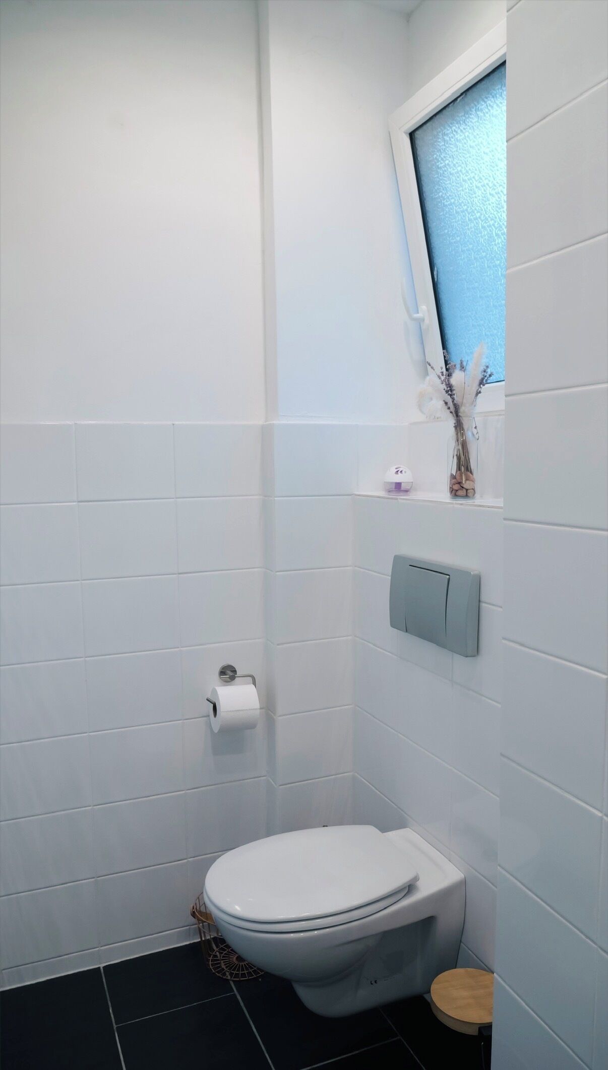 Un baño pequeño y moderno con azulejos blancos, un inodoro, un estante con artículos de aseo y una ventana con un tono azulado.