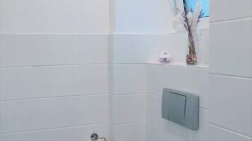 Un baño pequeño y moderno con azulejos blancos, un inodoro, un estante con artículos de aseo y una ventana con un tono azulado.