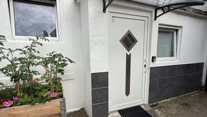Une porte blanche ornée d’une fenêtre en forme de losange, une petite fenêtre avec des stores et un auvent métallique au-dessus de la porte. On y trouve des jardinières en bois avec des fleurs rouges et une grille métallique au sol.