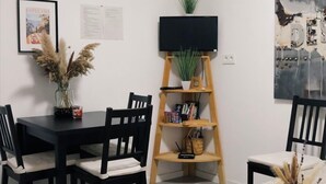 Un coin repas avec une table et des chaises noires, un buffet en bois agrémenté d’une plante, un téléviseur fixé au mur et un tableau encadré.