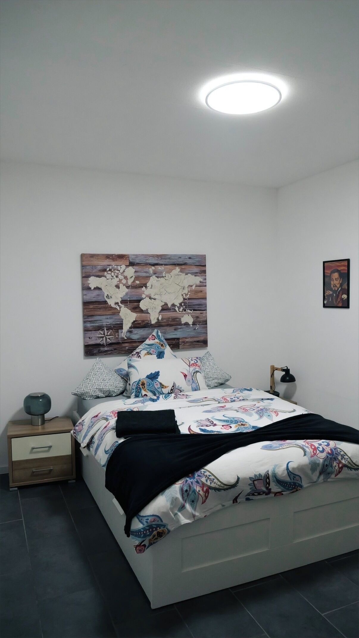 A bedroom with a bed, a nightstand, a lamp, a world map artwork, and a framed picture on the wall.