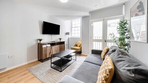 Ett modernt vardagsrum med en grå soffa, ett svart soffbord, ett träskåp, en platt-TV och ett stort fönster med utsikt över omgivningen.