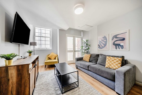 Un salon moderne avec un canapé gris, un bureau en bois et un téléviseur à écran plat. Des tableaux abstraits ornent le mur et une plante est posée sur le bureau.