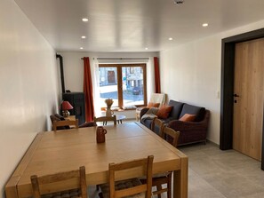 Een gezellige woonkamer met een houten eettafel, een bank met kussens, een open haard en uitzicht door een groot raam.