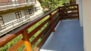 A balcony with a wooden railing overlooking a garden and a building with multiple windows and a red roof.
