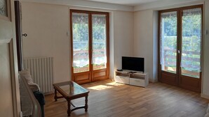 A spacious living room with a wooden floor, a glass-top coffee table, a flat-screen TV on a white cabinet, and large sliding glass doors with floral curtains.