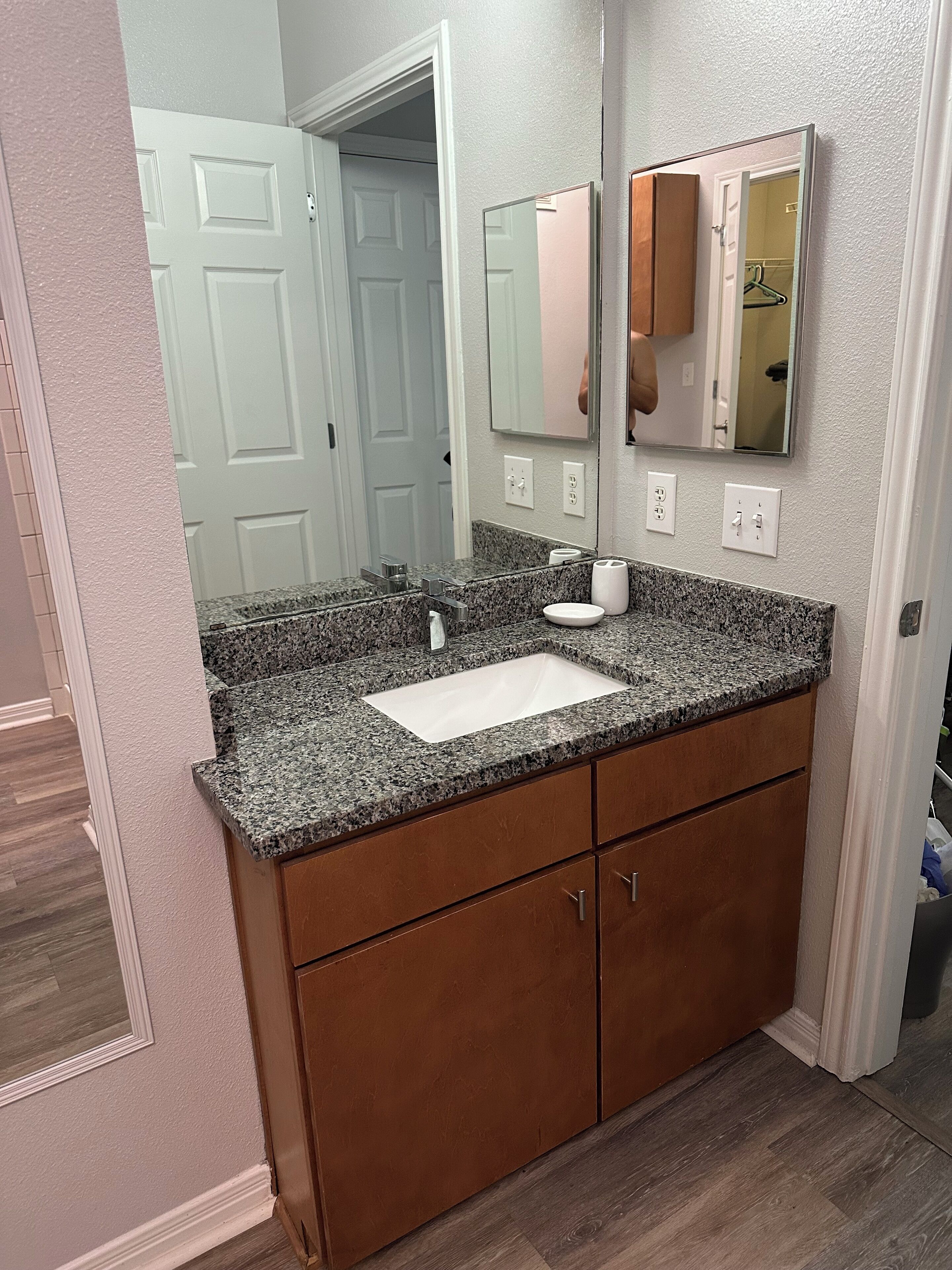 A bathroom with a granite countertop sink, wooden cabinets, a mirror, and a door leading to another room.