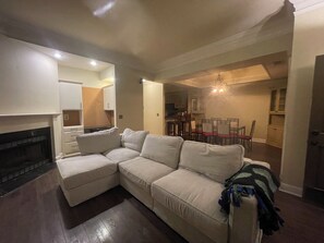 Ein Wohnzimmer mit Ecksofa, Kamin, Essbereich mit einer Pendelleuchte und einer dahinterliegenden Küche.