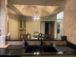 Eine Küche mit schwarzem Arbeitsplatz, einem Spülbecken und einem Spiegel, der einen Essbereich mit einer Kandelaberbeleuchtung reflektiert.