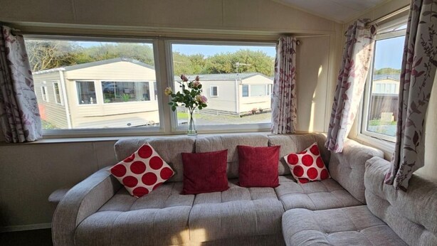 A sofa with four red pillows and a vase with flowers on a windowsill. - Willerby Seasons: 3BR Caravan (Morecambe)