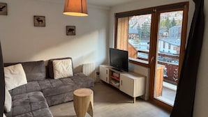 Ein modernes Wohnzimmer mit einer grauen Eckcouch, einem hölzernen Hocker, einem Flachbildfernseher auf einem weißen Schrank und einem großen Fenster mit Blick auf Gebäude und Bäume.