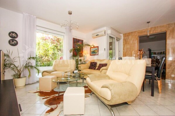 Ein modernes Wohnzimmer mit einem beigen Sofa, einem Glastisch und einem Essbereich mit Holztisch und Stühlen. Üchtige Pflanzen und ein großes Fenster sorgen für natürliches Licht.