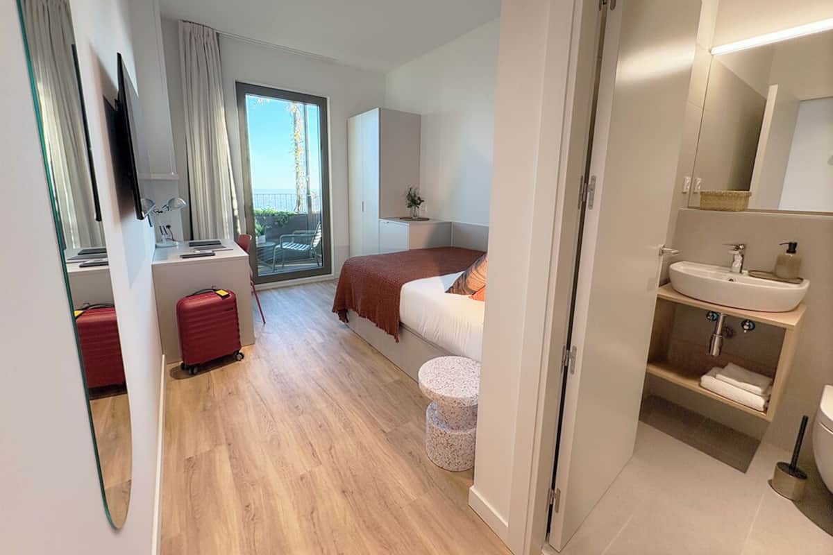A modern hotel room with a bed, a desk, a bathroom, and a view of the outdoors.