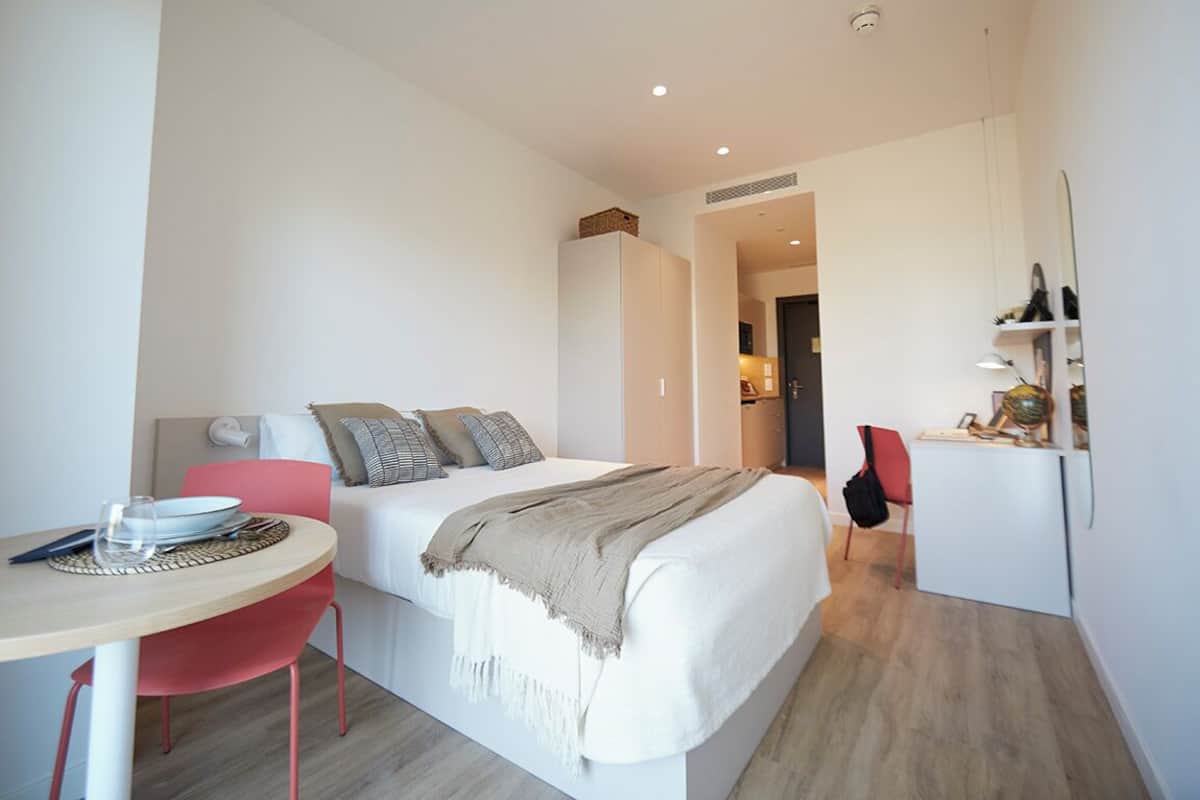 A modern hotel room with a bed, a desk, a chair, and a small kitchenette.