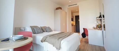 A modern hotel room with a bed, a desk, a chair, and a small kitchenette.