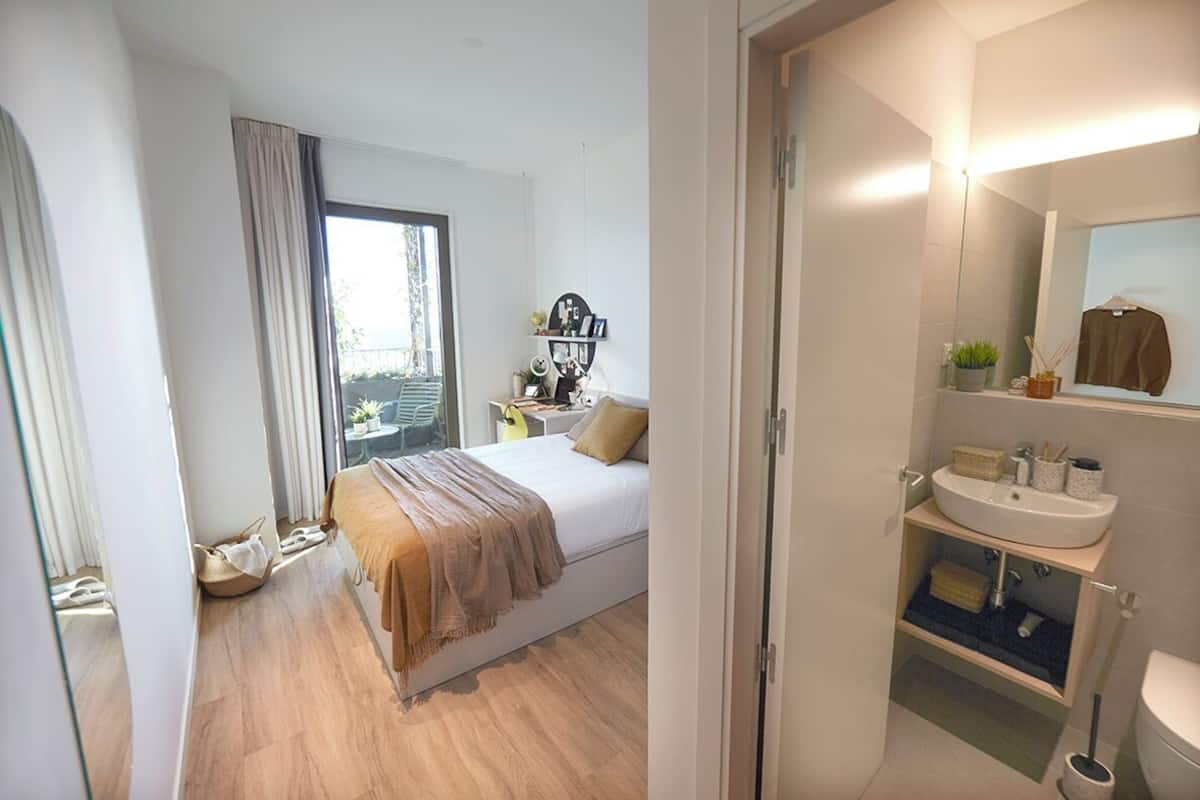 A bedroom with a bed, a window with curtains, a bedside table with a lamp, and a bathroom with a sink and mirror.