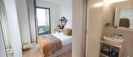 A bedroom with a bed, a window with curtains, a bedside table with a lamp, and a bathroom with a sink and mirror.