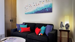 Een moderne woonkamer met een zwarte bank, kleurrijke kussens, een ronde salontafel en een abstract schilderij aan de muur.