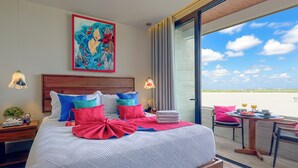 Een moderne slaapkamer met een groot bed, kleurrijke kussens, een schilderij aan de muur en een balkon met een tafel en stoelen.