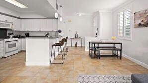Een moderne keuken met witte kastjes, een centraal eiland en barstoelen. Er is een eethoek met een tafel en stoelen, en een raam met jaloezieën.