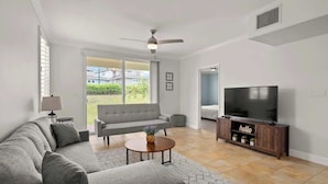Een moderne woonkamer met een grijze hoekbank, een ronde salontafel, een televisie op een houten dressoir en een plafondventilator.