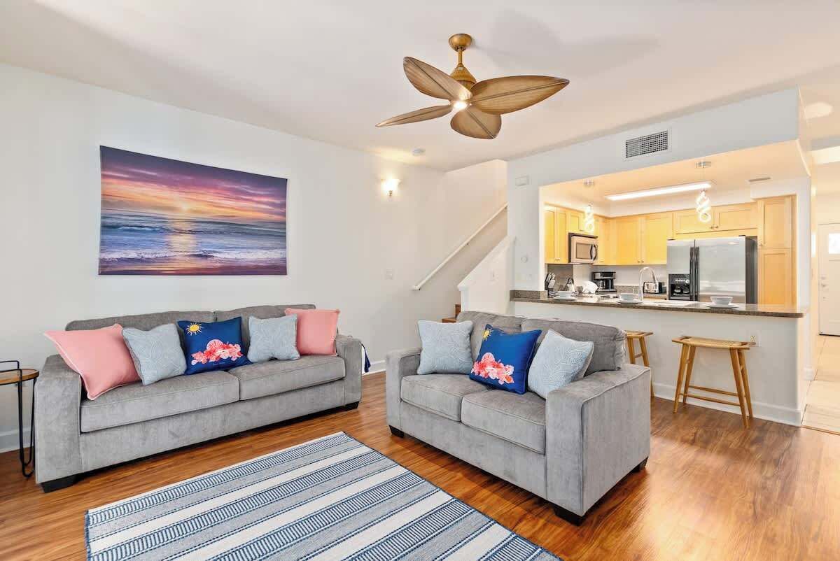 A modern living room with two grey sofas, a wooden floor, a ceiling fan, and a kitchen visible through an open door.