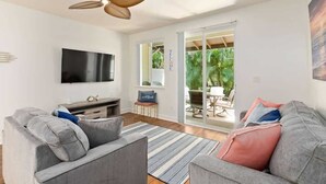 A modern living room with a flat-screen TV, grey sofas, a ceiling fan, and a view through glass doors to an outdoor patio. - Luxury Oasis Resort Villa (Kapolei)