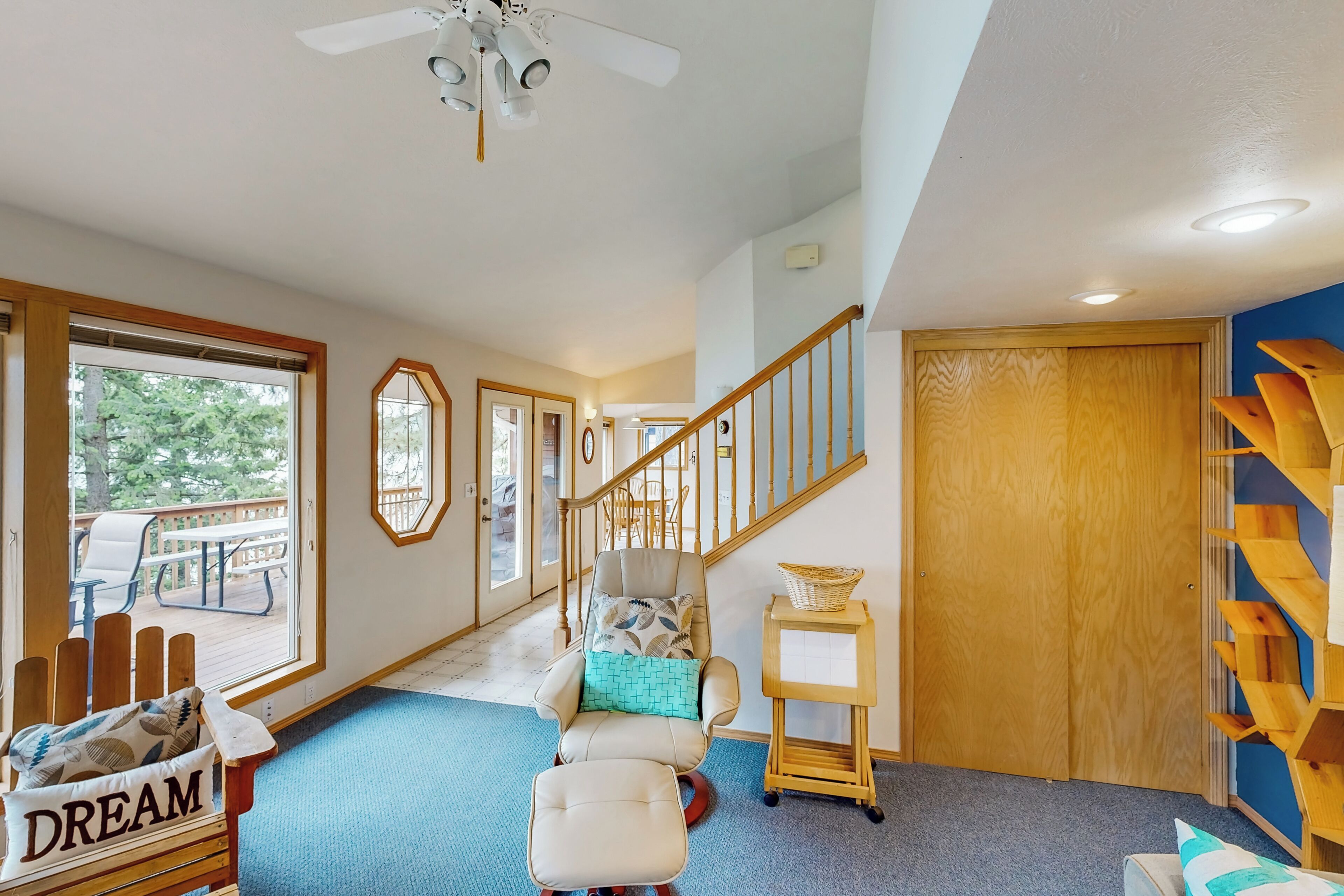 Un intérieur chaleureux avec un banc en bois portant l’inscription « RÊVE », un fauteuil rembourré avec un coussin bleu, une petite table avec un panier et un escalier menant à une porte en bois.