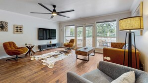 A modern living room with hardwood flooring, a ceiling fan, a flat-screen TV, a sofa, two armchairs, a coffee table, and a large window with a view of the outdoors. - ‘Lakeside Getaway’ w/ Patio on Lady Bird Lake (Austin)