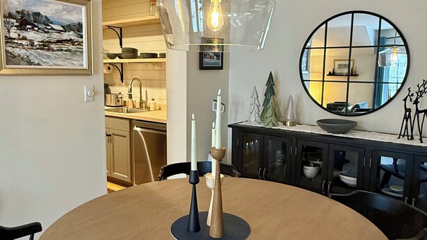 A round dining table with a central light fixture, surrounded by chairs. A kitchen area is visible in the background, featuring a sink and cabinets. - Quiet Village Gem! Cozy retreat for two. (Stowe)