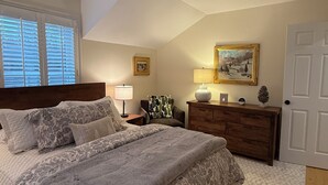 A bedroom with a large bed, a dresser, a nightstand, a lamp, a framed picture, and a door. - Quiet Village Gem! Cozy retreat for two. (Stowe)