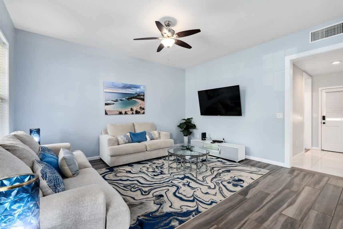 Een moderne woonkamer met een beige bank, een glazen salontafel, een tapijt met een patroon, een plafondventilator en een flatscreen televisie aan de muur.