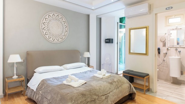 Habitación de hotel con cama, lámparas de noche, mesitas de noche, un banco, un espejo y un baño con lavamanos y espejo.