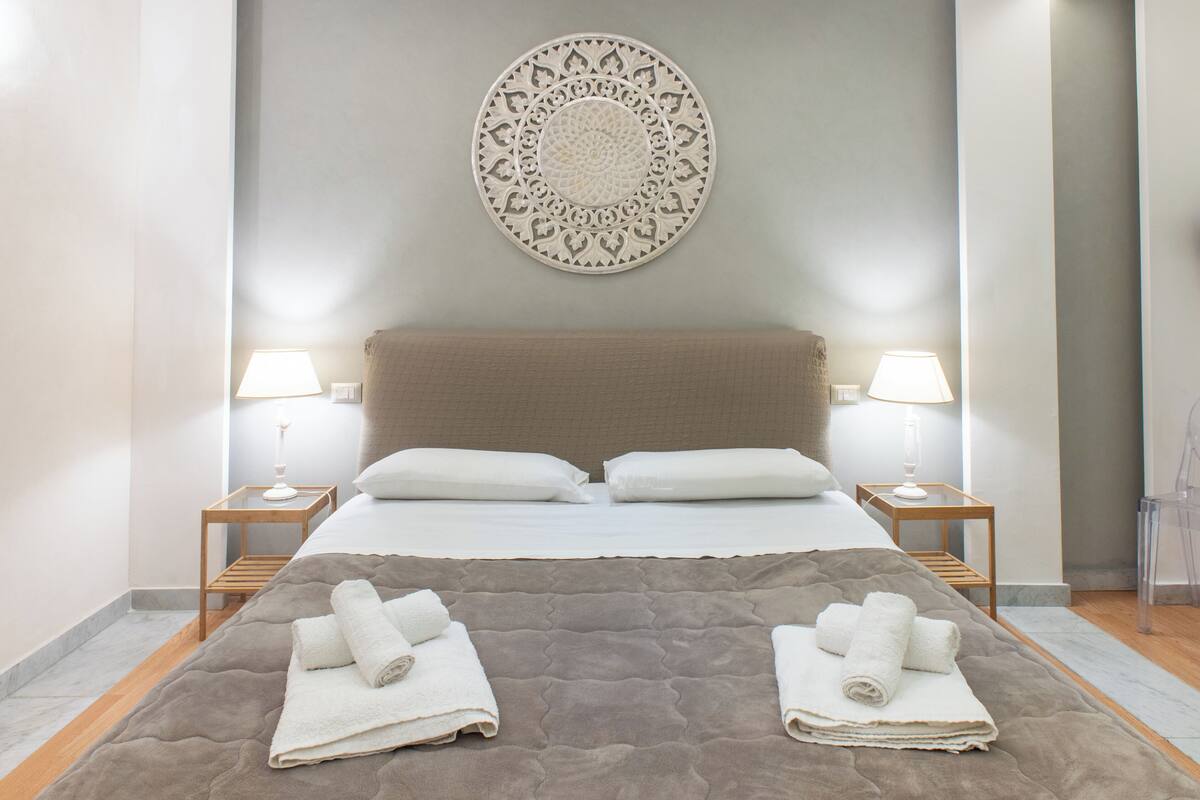A hotel room with a bed, two bedside tables with lamps, a wall-mounted decorative plate, and two rolled towels on the bed.