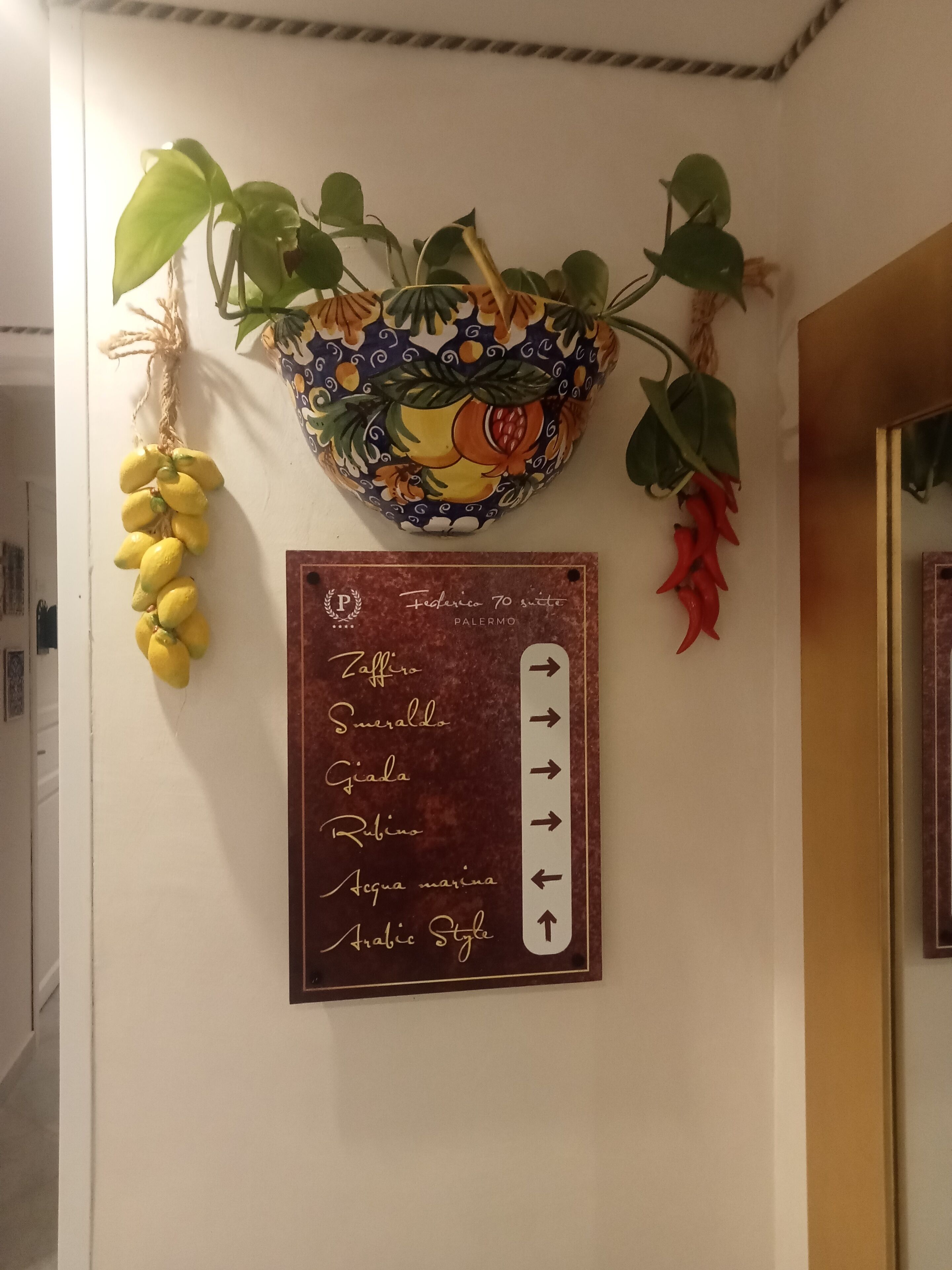 A wall with a decorative hanging featuring lemons, chili peppers, and a bowl with a fruit pattern. Below, a sign lists room names and directions.