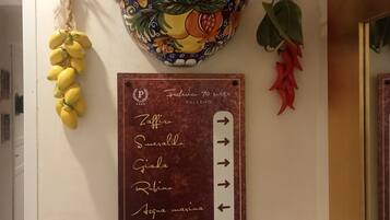 A wall with a decorative hanging featuring lemons, chili peppers, and a bowl with a fruit pattern. Below, a sign lists room names and directions.