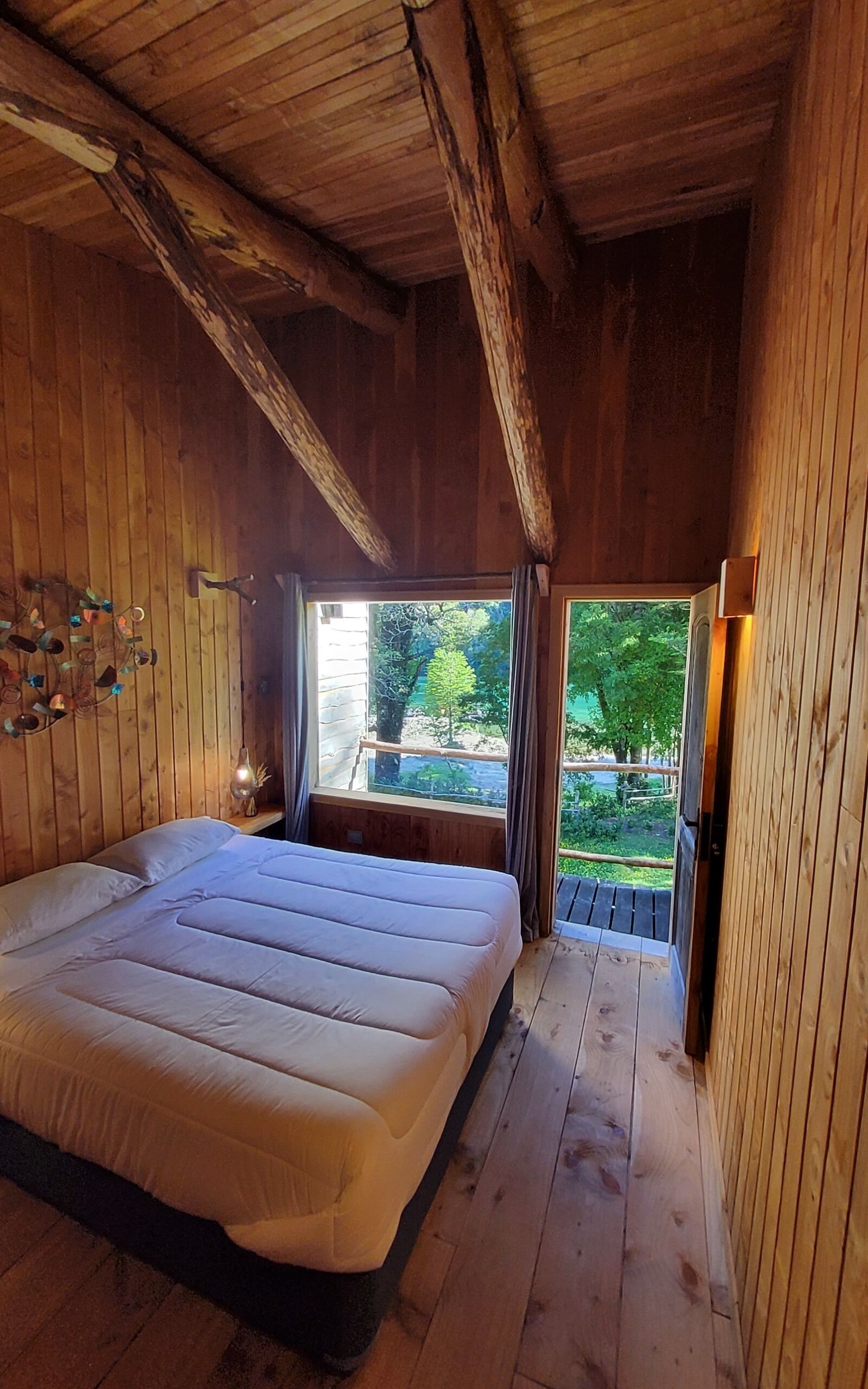 Un dormitorio de cabaña de madera con una cama grande, una ventana con cortinas y vista a la vegetación.