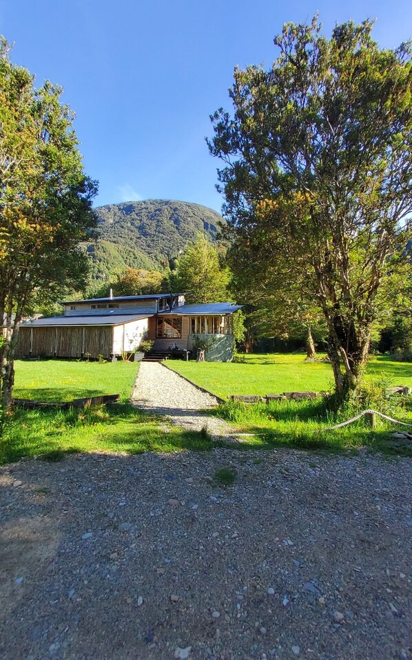 A rustic cabin with a gravel driveway leading to a wooden deck, surrounded by lush greenery and a mountain backdrop. - Salvatierra Lodge (Cisnes)