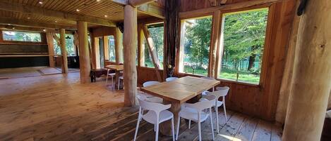 A spacious wooden dining area with a large table and chairs, surrounded by large windows offering views of a wooded area.