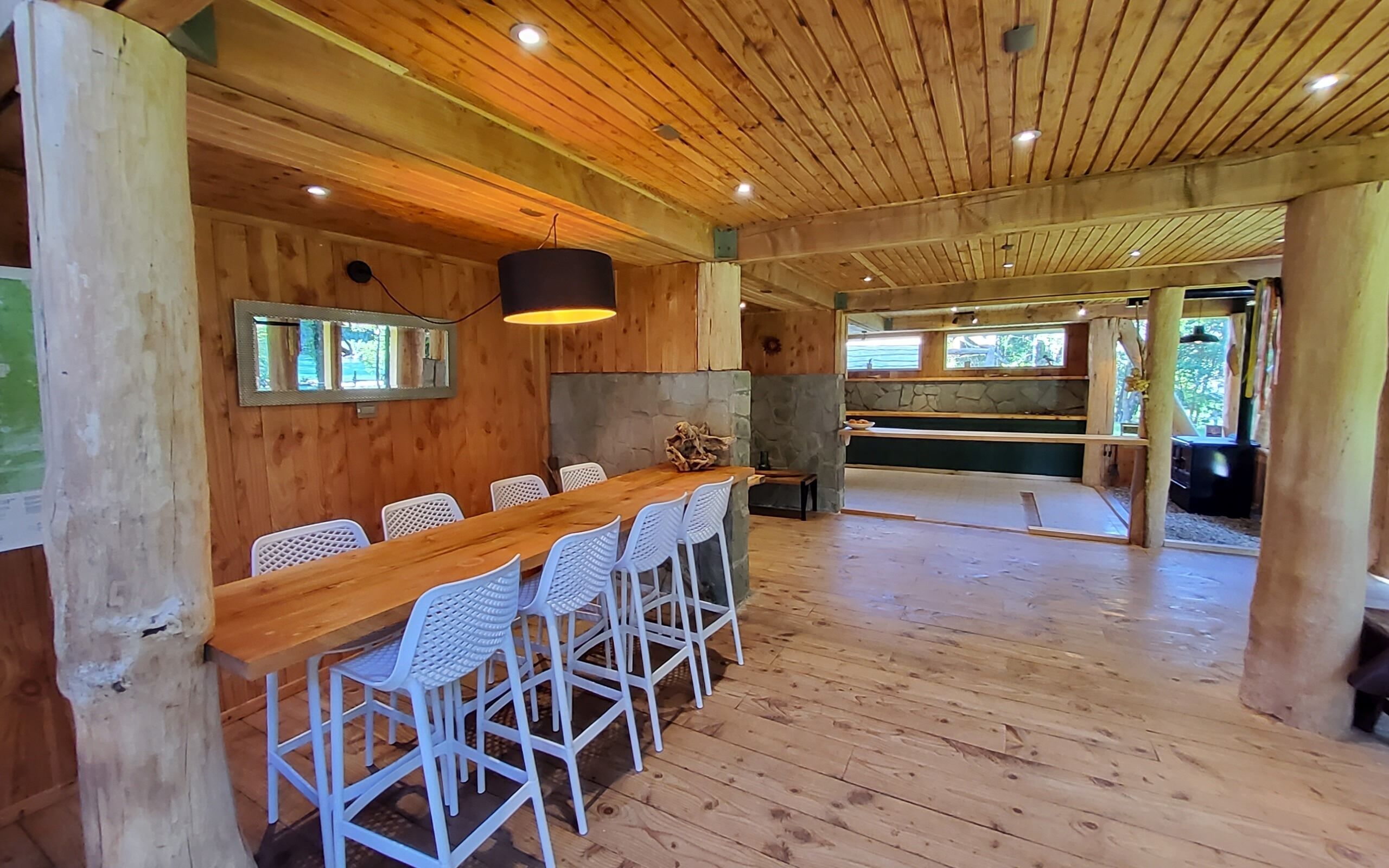 A wooden interior with a long table and white chairs, a stone wall, and a view of the outdoors.