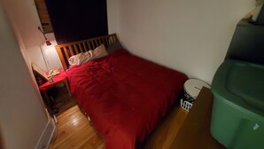 Ein kleiner, schwach beleuchteter Raum mit einem Einzelbett, einem Nachttisch, einer Lampe und einer grünen Mülltonne.