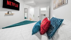 Un dormitori modern amb un llit gran, una televisió penjada a la paret que mostra 'NETFLIX', una escala que mena a una altra habitació i un quadre a la paret.