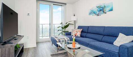 A modern living room with a large flat-screen TV, a blue sofa, a glass coffee table, and a view through a large sliding glass door.