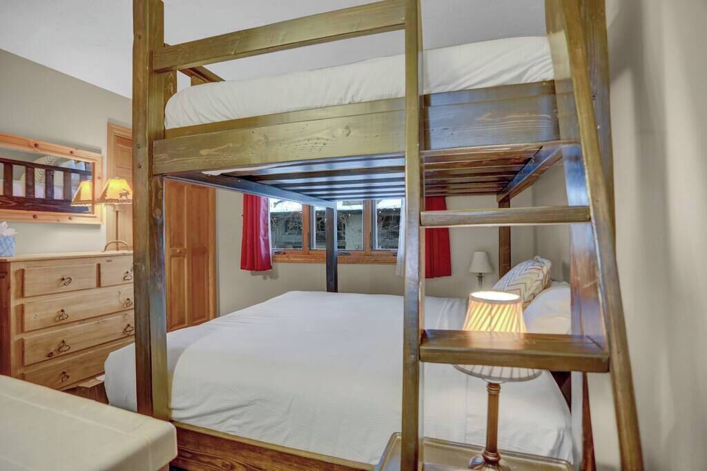 A bunk bed room with a wooden ladder, a dresser, a lamp, and a window with red curtains.