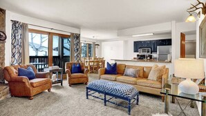 Ein geräumiges Wohnzimmer mit einer Couch, Sesseln und einem Couchtisch. Im Hintergrund ist eine Küche mit modernen Geräten zu sehen.